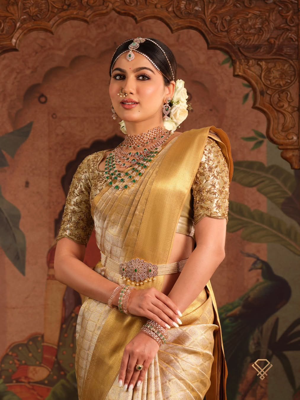 A Meralda muse in a gold tissue Kanjivaram saree against a hand-painted peacock mural, wearing layered antique-gold and polki jewellery